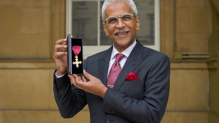 Professor Mahendra Patel smiling happily while holding up his OBE.